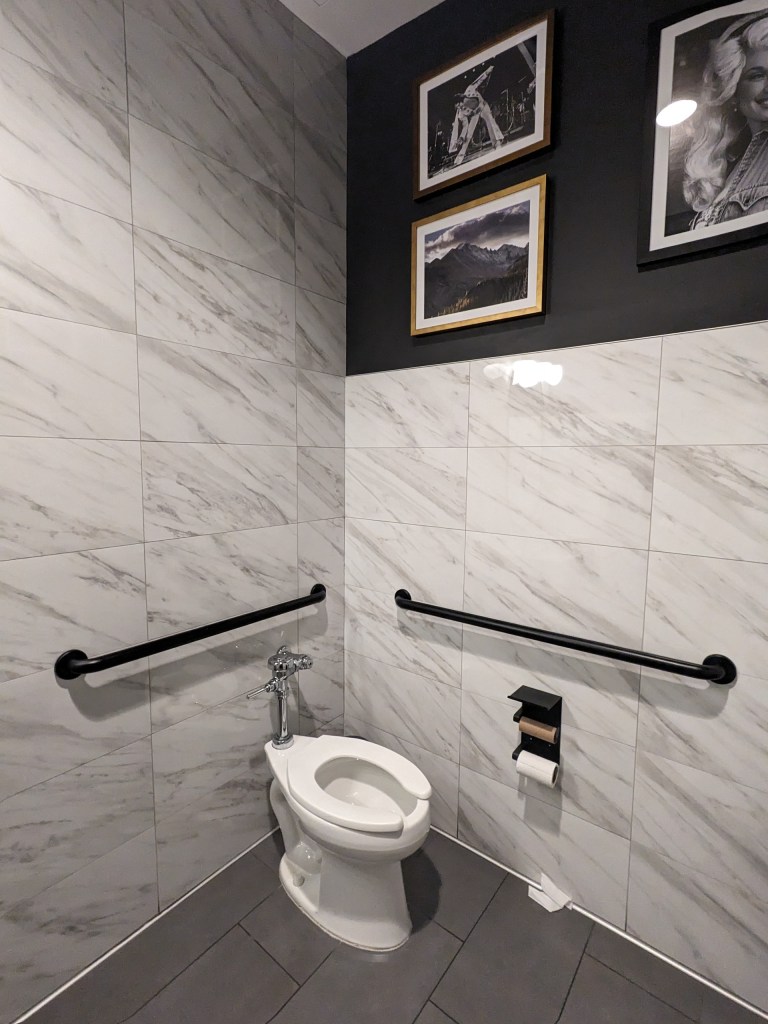 An accessible bathroom corner showing a white toilet with black horizontal grab bars on both adjacent tiled walls. The upper wall is painted dark grey and decorated with framed black-and-white photographs.