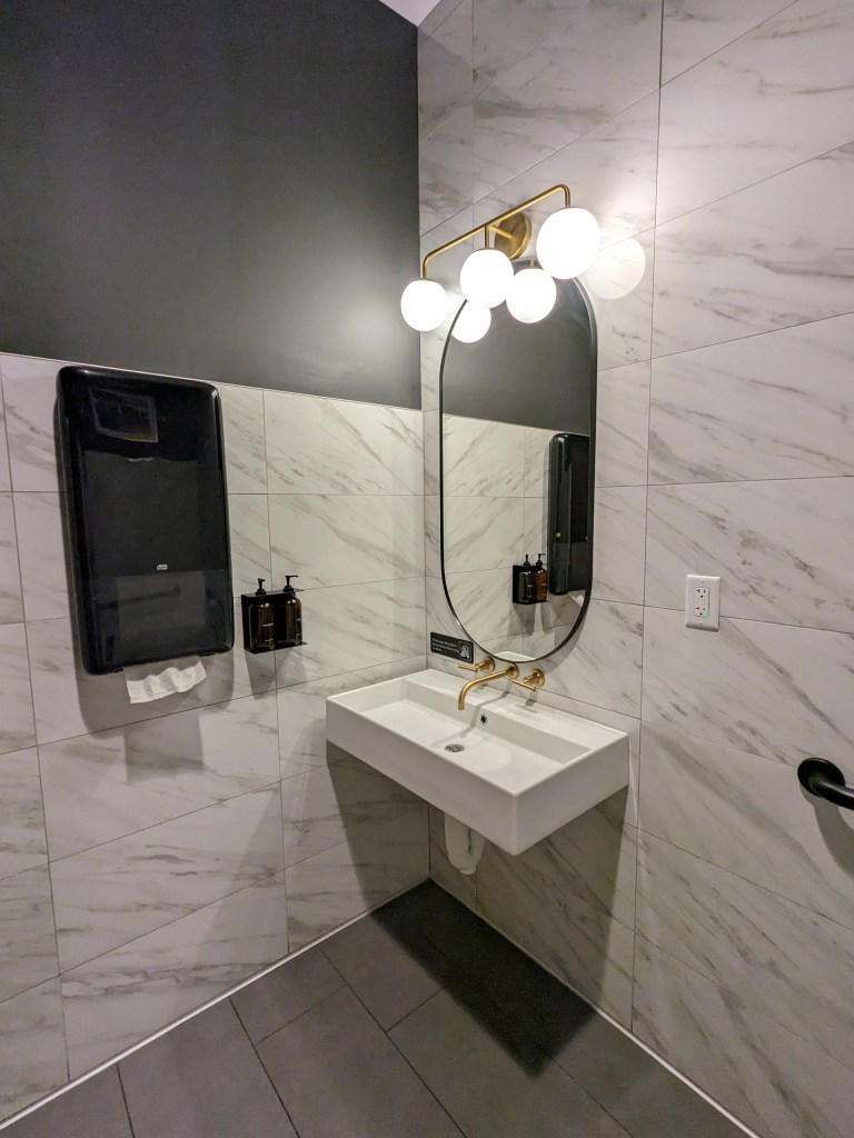 A modern accessible bathroom with a wall-mounted rectangular sink beneath an oval mirror, illuminated by a brass light fixture with globe bulbs. The walls are tiled in white marble effect with dark grout, and a paper towel dispenser and soap bottles are mounted beside the sink.